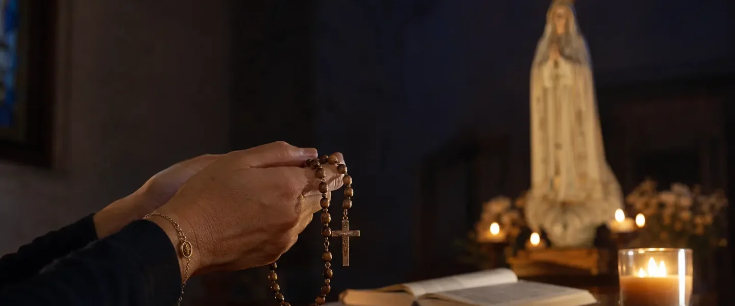 Mani di una fedele che pregano il Rosario davanti alla Madonna di Fatima, accanto a una candela accesa